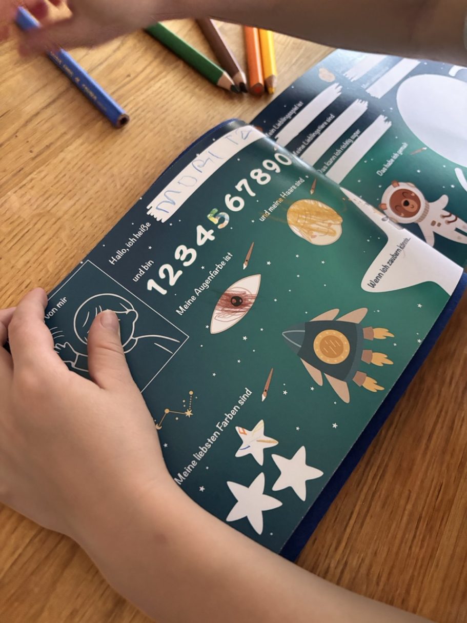Freundebuch Weltall Eine Hand hält ein Buch mit bunten Zeichnungen von Planeten, Sternen und Zahlen.