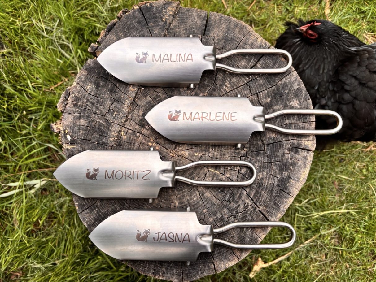 personalisierter Mini-Klappspaten Vier Metallspaten mit Griffen auf einem Baumstamm, im Hintergrund ein Huhn.
