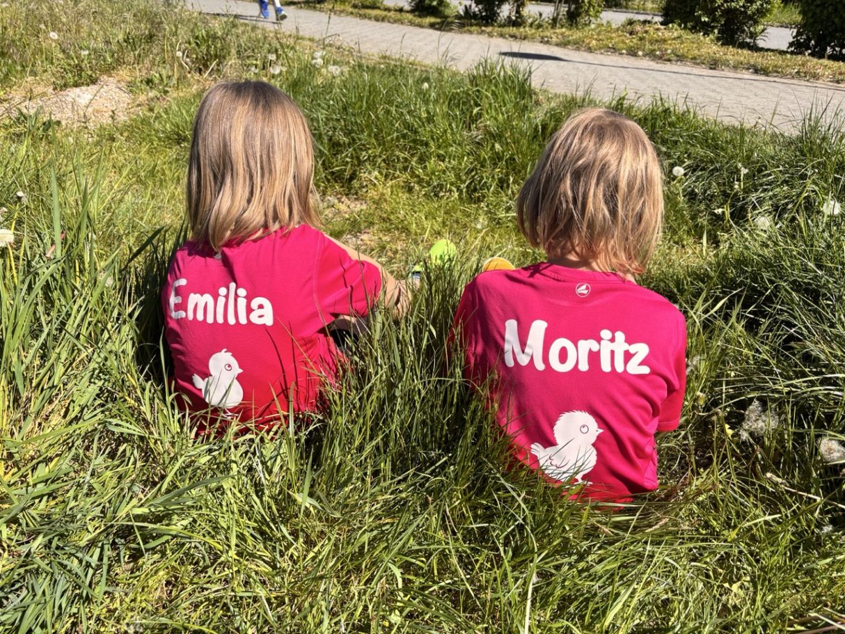 Trikot mit Namen und Bild Zwei Kinder sitzen im Gras, tragen rote T-Shirts mit den Namen Emilia und Moritz.